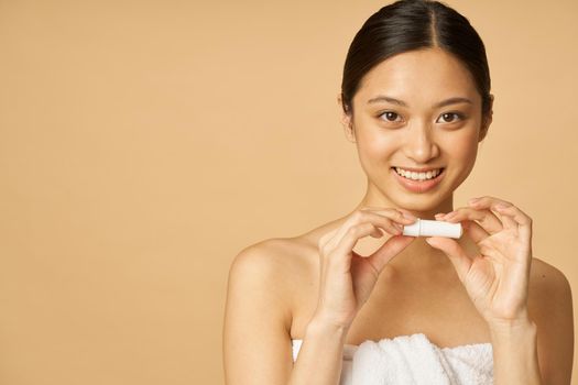 Portrait of adorable young woman wrapped in towel smiling while holding lip balm, posing isolated over beige background. Beauty treatment concept