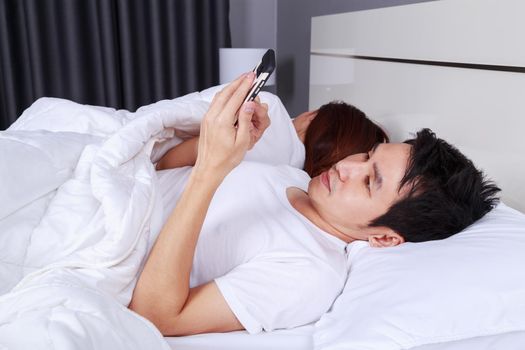 young man using his mobile phone in bed while his wife is sleeping next to him
