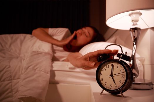 sleepy young woman in bed with eyes closed extending hand to alarm clock in the morning