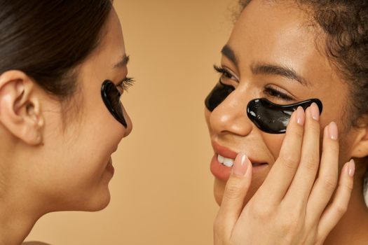 Close up portrait of lovely young women looking at each other, applying black hydro gel under eye patches on facial skin, posing isolated over beige background. Skincare routine, diversity concept