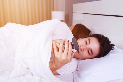 young man using his mobile phone in bed while his wife is sleeping next to him
