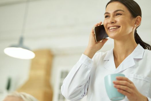 Happy nurse holding cup of coffee and talking on the mobile phone while standing in room