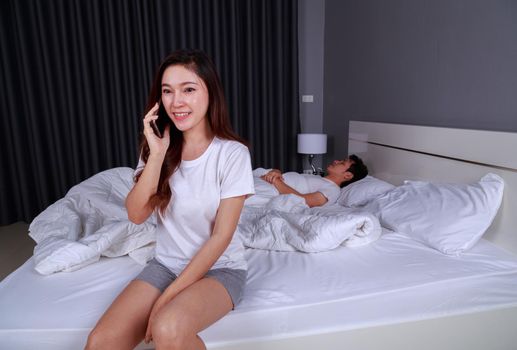 young happy woman talking on mobile phone in bed while her husband is sleeping next to her
