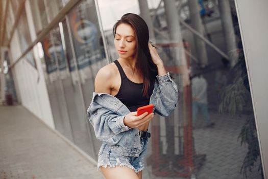 Beautiful girl in a jeans jacket. Woman standing near large building. Brunette use the phone