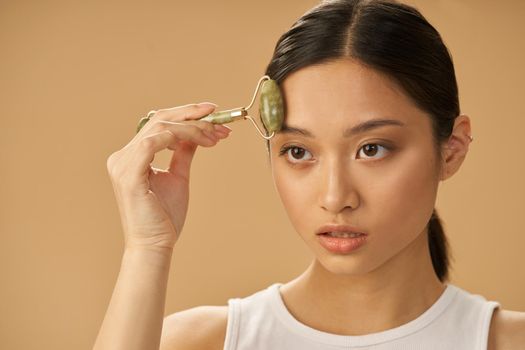 Attractive young woman looking aside while using jade roller for massaging her face, posing isolated over beige background. Skincare concept