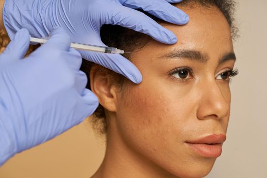 Portrait of beautiful young woman looking away while getting face injections isolated over beige background. Beauty, cosmetology concept