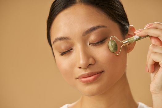 Portrait of relaxed young woman using jade roller for massaging her face, posing with eyes closed isolated over beige background. Skincare concept