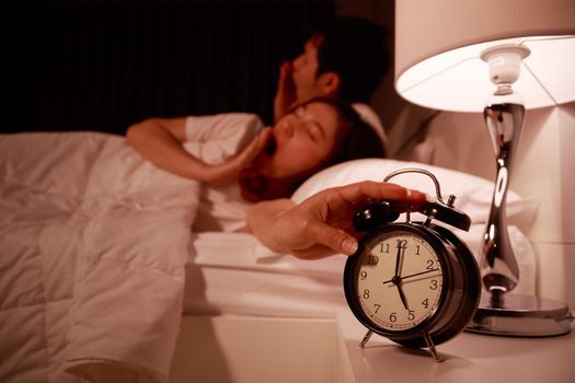 sleepy young couple in bed with extending hand to alarm clock in the morning