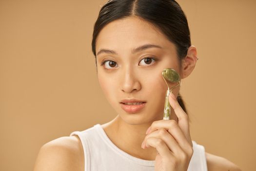 Facial beauty treatment. Beautiful young woman looking at camera while getting massage face using jade roller isolated over beige background. Skincare concept