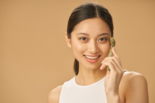 Cheerful beautiful young woman smiling at camera while using jade roller for massaging her face, posing isolated over beige background. Skincare concept