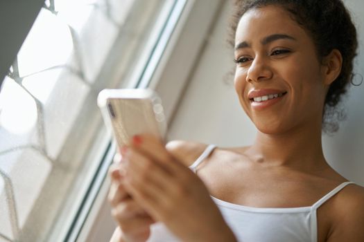 Portrait of cheerful young mixed race woman smiling, using smartphone, feeling cozy while sitting by the window at home. Lifestyle, leisure concept