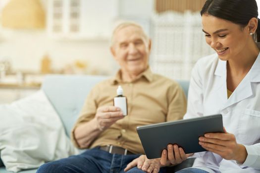 Happy nurse using tablet while talking with aged man with with medicine in hand in the room