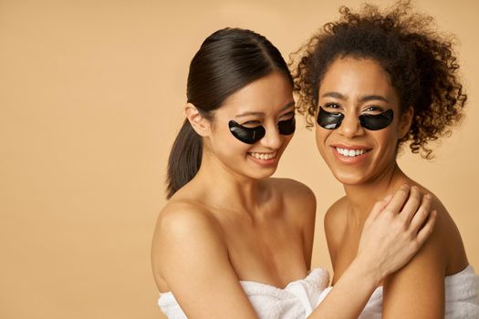 Smiling young women wrapped in towel posing with applied black hydro gel under eye patches isolated over beige background. Skincare routine concept