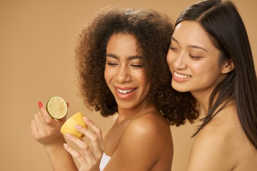 Two lovely mixed race young women looking happy, holding lemon and lime cut in half while posing together isolated over beige background. Health and beauty concept