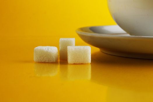 A white cup on a saucer with tea or coffee next to lie cubes of sugar slices and a doughnut in the glaze on a bright yellow background with a place for a copyspace. Still life with sweet food.