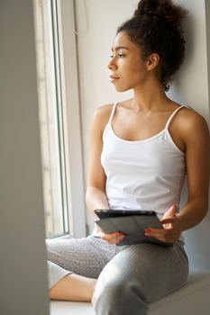 Thoughtful young woman with tablet pc looking out the window, feeling cozy while sitting alone by the window at home. Lifestyle, weekend concept