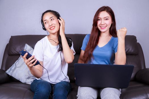 two young woman using laptop computer and listening to music in headphones with moblie on sofa at home