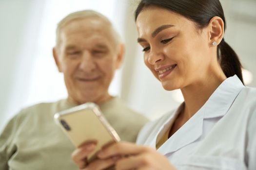 Close up of happy aged man and female doctor in white coat standing and using mobile phone while reading message