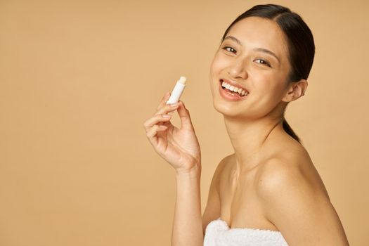 Portrait of beautiful young woman wrapped in towel smiling at camera while holding lip balm, posing isolated over beige background. Beauty treatment concept