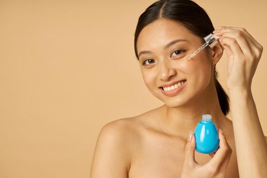 Portrait of charming young woman smiling at camera, dropping oil serum with pipette on face for perfect skin, posing isolated over beige background. Beauty treatment concept