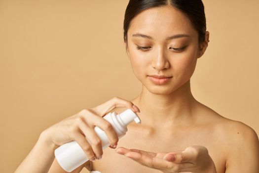 Studio portrait of natural young woman holding a bottle of gentle foam facial cleanser isolated over beige background. Beauty products and skin care concept