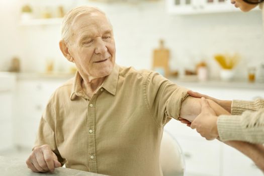 Serious aged man sitting at the table while having medical doctor touch his arm