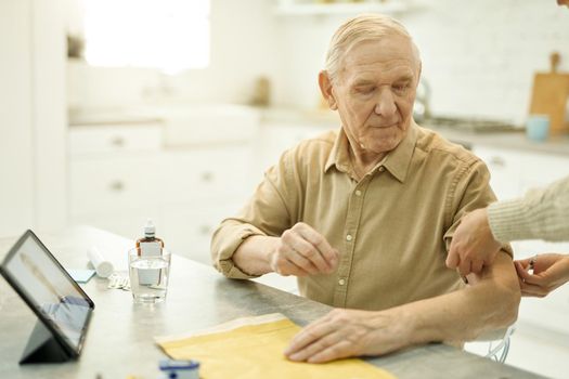 Calm aged man sitting at the table while medical doctor rolling up his sleeve