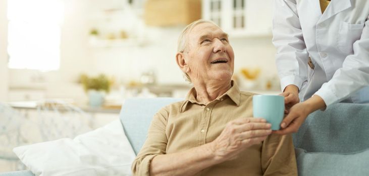 Female doctor in white coat serving a cup to senior citizen at his living room while helping him