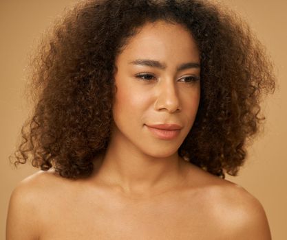 Portrait of lovely mixed race young woman with curly hair looking aside posing isolated over beige background. Female beauty portrait