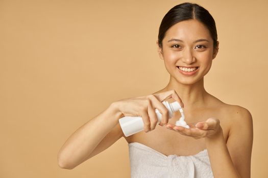 Studio portrait of pretty young woman smiling at camera, holding a bottle of gentle foam facial cleanser isolated over beige background. Beauty products and skin care concept. Front view