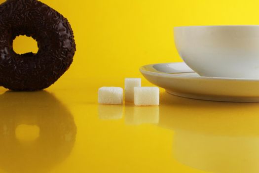 A white cup on a saucer with tea or coffee next to lie cubes of sugar slices and a doughnut in the glaze on a bright yellow background with a place for a copyspace. Still life with sweet food.