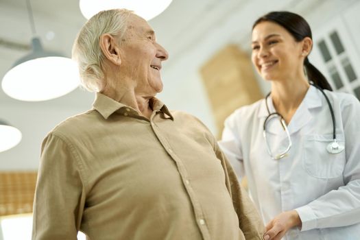 Pleased elderly man looking at a woman in white lab coat visiting him at home