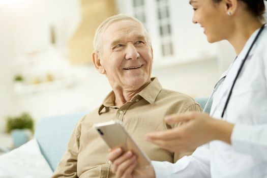 Medical doctor holding smartphone while talking to senior man at his home