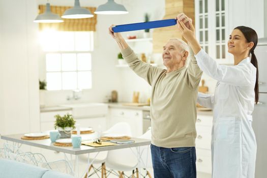 Aged man raising his hands up and doing exercises with fitness rubber band, female doctor helping him