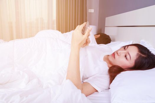 young woman using her mobile phone in bed while her husband is sleeping next to her
