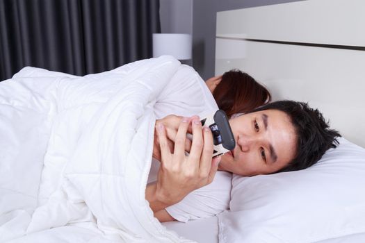 young man using his mobile phone in bed while his wife is sleeping next to him
