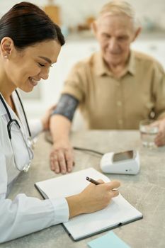 Cheerful female medic writing notes while sitting beside a senior gentleman with manometer on the table