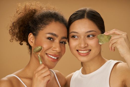 Portrait of lovely young women smiling while using jade roller and facial gua sha, posing together isolated over beige background. Skincare, diversity concept