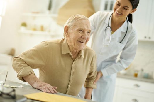Friendly medical worker helping senior citizen get up from a chair while visiting him at home