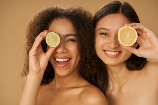 Portrait of two joyful mixed race young women looking excited, covering eyes with lemon and lime cut in half while posing together isolated over beige background. Health and beauty concept