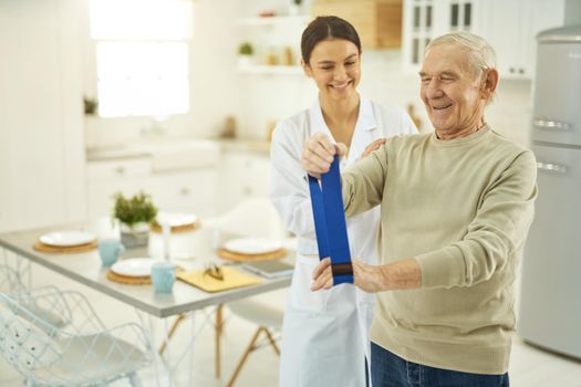 Smiling pensioner doing exercises and holding a fitness elastic band in hands while talking to nurse