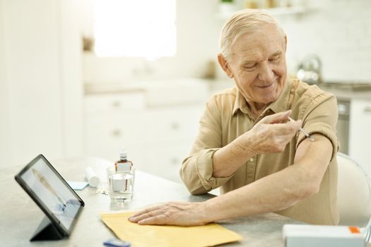 Joyous senior man sitting with rolled-up sleeve and giving himself a shoulder injection