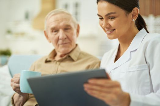 Smiling female doctor using tablet while talking with old man with cup of hot drink in the room