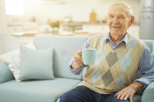 Smiling male pensioner sitting on couch in living room while holding cup of hot drink. Copy space