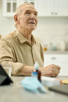 Concentrated aged man sitting at the table with some medication on it and looking up