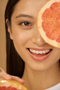 Close up portrait of joyful mixed race young woman smiling at camera, covering one eye with grapefruit cut in half, posing isolated over beige background. Health and beauty concept