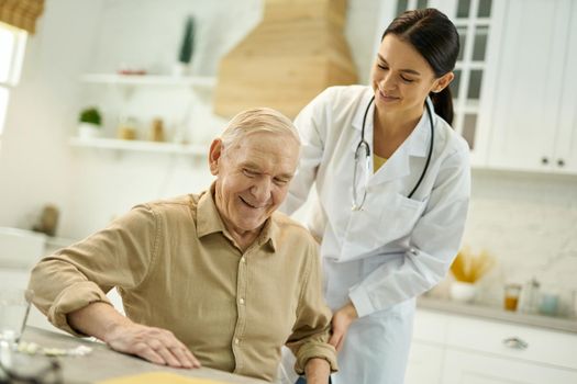 Positive elderly man smiling while getting up from the chair with the help of attentive young doctor