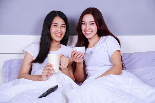 two best friends watching tv with remote and drinking a cup of coffee on bed in the bedroom 