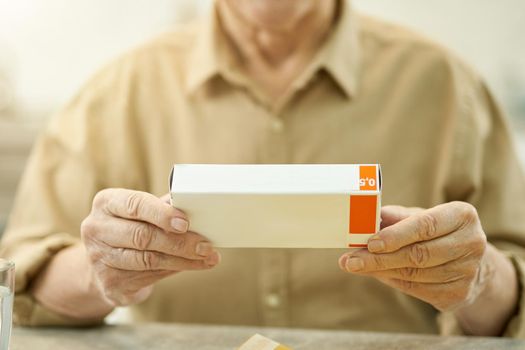 Copy space photo of senior man in button-up shirt holding a blank box of medication while sat at the table