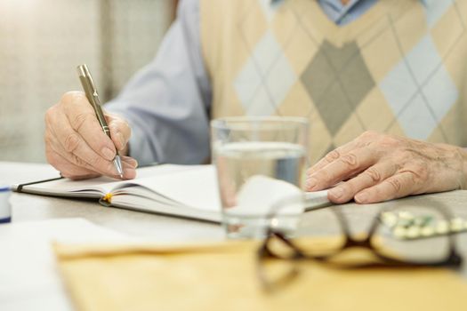 Cropped photo of an aged man writing in a notebook while sitting at the table